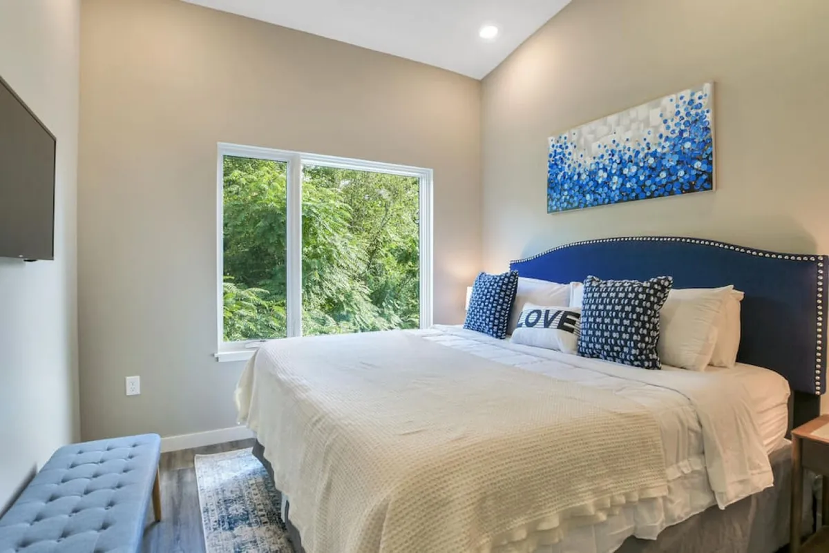 A king bedroom featuring a bed with decorative pillows, a large window overlooking greenery, colorful artwork above the bed, and a TV mounted on the wall. The room is airy and infused with natural light, offering a tranquil retreat.