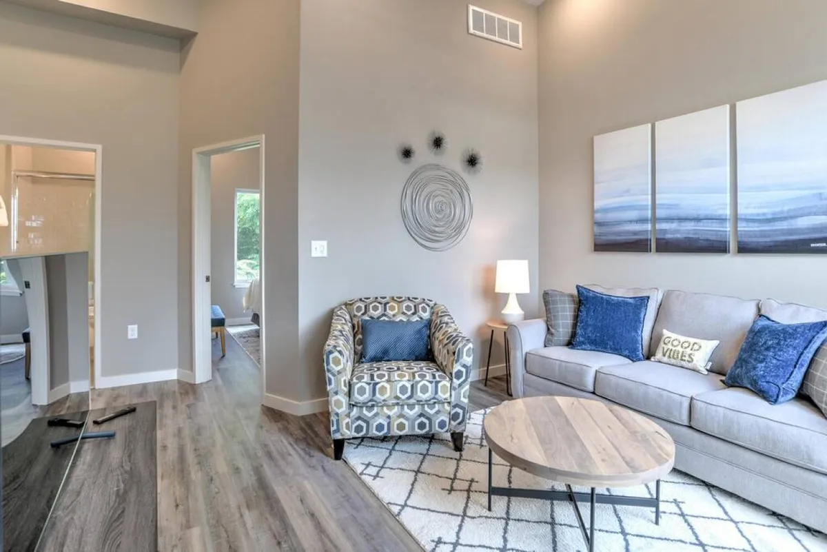 A cozy, modern living area featuring a textured wall art, a patterned armchair, and a round coffee table, all bathed in natural sunlight. The space is both inviting and relaxing, suggesting a comfortable retreat after a day out.