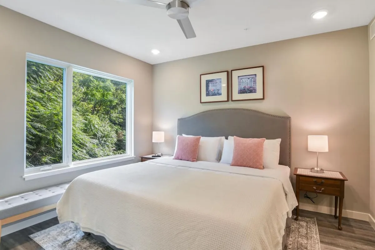 A king bedroom with a bed dressed in white linens and pink accent pillows, large windows providing a view of lush greenery, and nightstands with lamps and curated artwork. The space is bathed in natural light, giving it a tranquil and inviting mood.