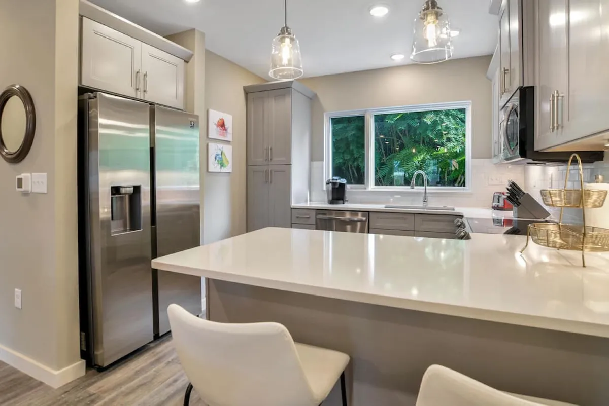 A contemporary kitchen with a stainless steel refrigerator, white quartz countertops, and a large window that fills the space with natural light and offers a view of greenery. The clean lines and modern finishes create a fresh, inviting setting.