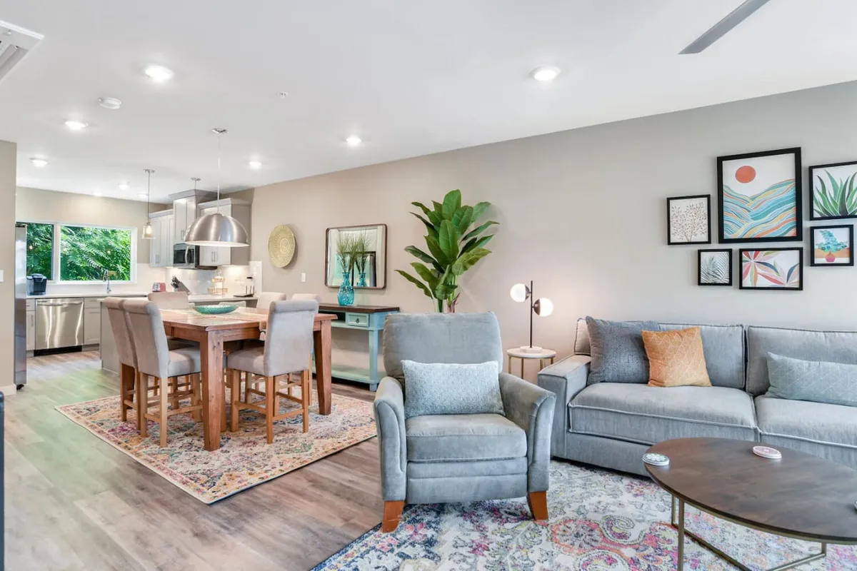 An open-plan gathering area featuring a modern dining table and chairs, a cozy sofa, striking wall art, and a lush, large plant. The space is accentuated by natural light and arranged for both dining and relaxing, reflecting a welcoming ambiance.